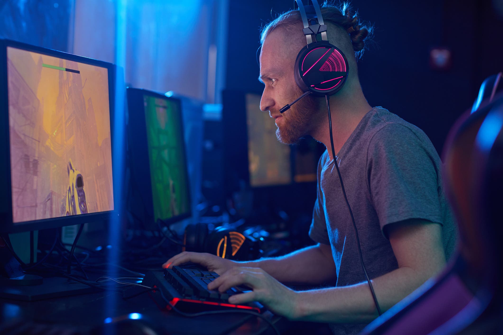 <p>Young bearded man in headphones looking at computer monitor and playing in computer game alone in dark room</p>
